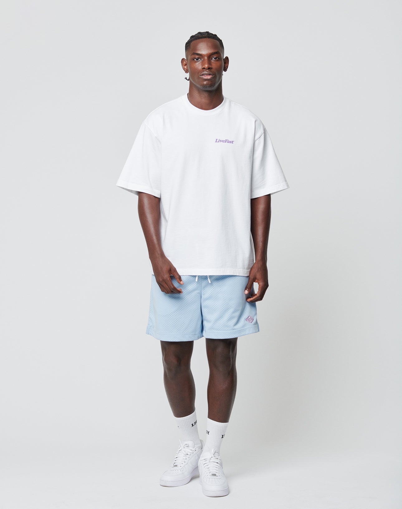 A man stands against a plain background, wearing a white oversized T-shirt and LIVE FAST DIE YOUNG Basic Mesh Shorts in light blue with an elastic waist, paired with white socks and sneakers. He faces forward with a neutral expression and relaxed posture.