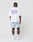 A person faces away, wearing a white LIVE FAST DIE YOUNG Basic 280 Tee with purple text, light blue shorts, white socks, and sneakers against a plain white background. The Oversize Fit tee is made from 100% Baumwolle (cotton).