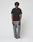 A person stands against a plain light background, wearing the LIVE FAST DIE YOUNG Live Fast Plain Tee in black (Regular Fit, 100% Baumwolle), paired with gray pants, white sneakers, and dark sunglasses. Their short curly hair and relaxed pose complete the look.