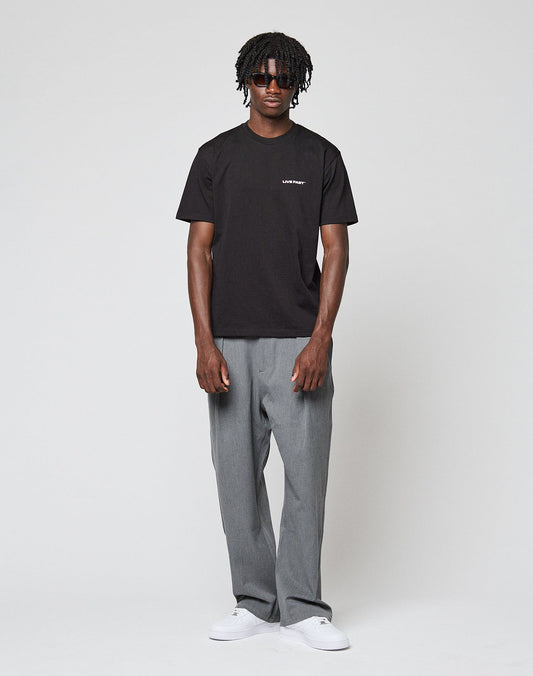 A person stands against a plain light background, wearing the LIVE FAST DIE YOUNG Live Fast Plain Tee in black (Regular Fit, 100% Baumwolle), paired with gray pants, white sneakers, and dark sunglasses. Their short curly hair and relaxed pose complete the look.