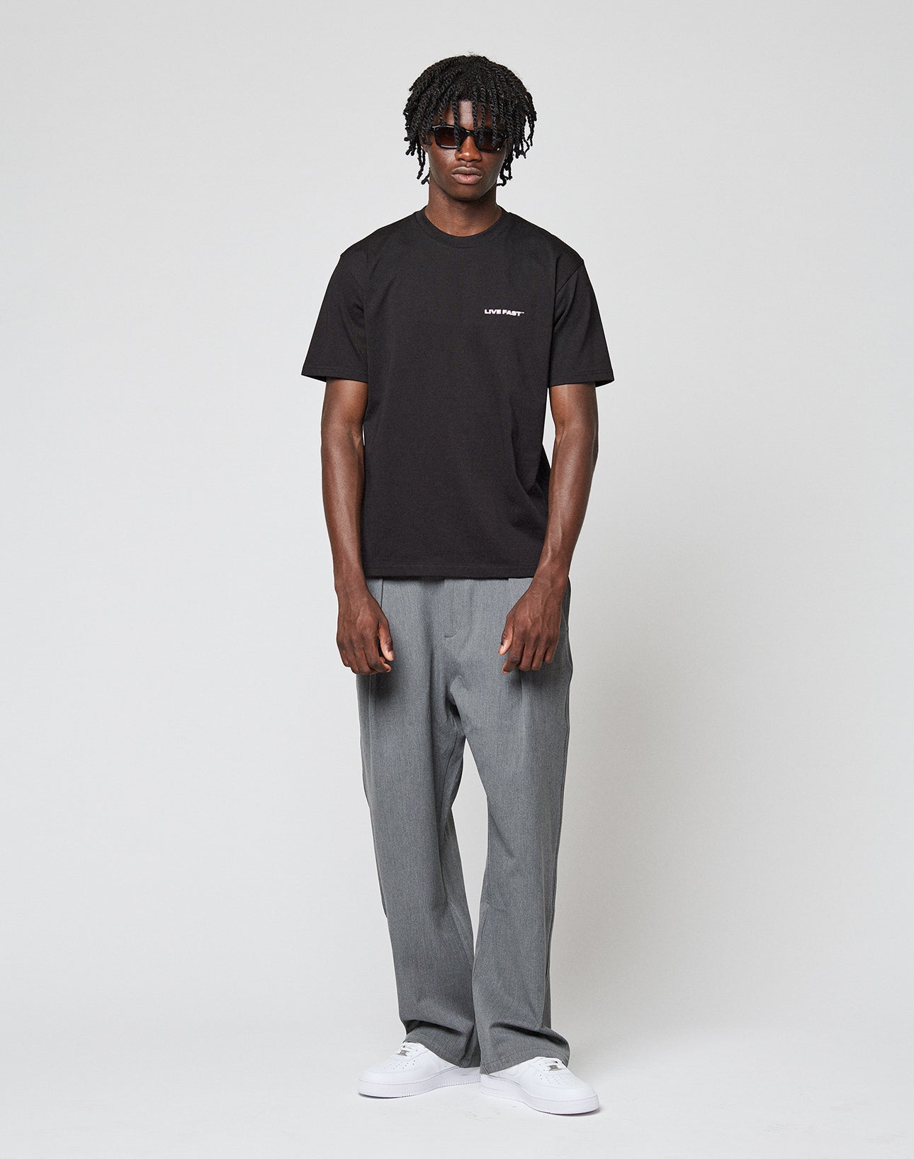 A person stands against a plain light background, wearing the LIVE FAST DIE YOUNG Live Fast Plain Tee in black (Regular Fit, 100% Baumwolle), paired with gray pants, white sneakers, and dark sunglasses. Their short curly hair and relaxed pose complete the look.