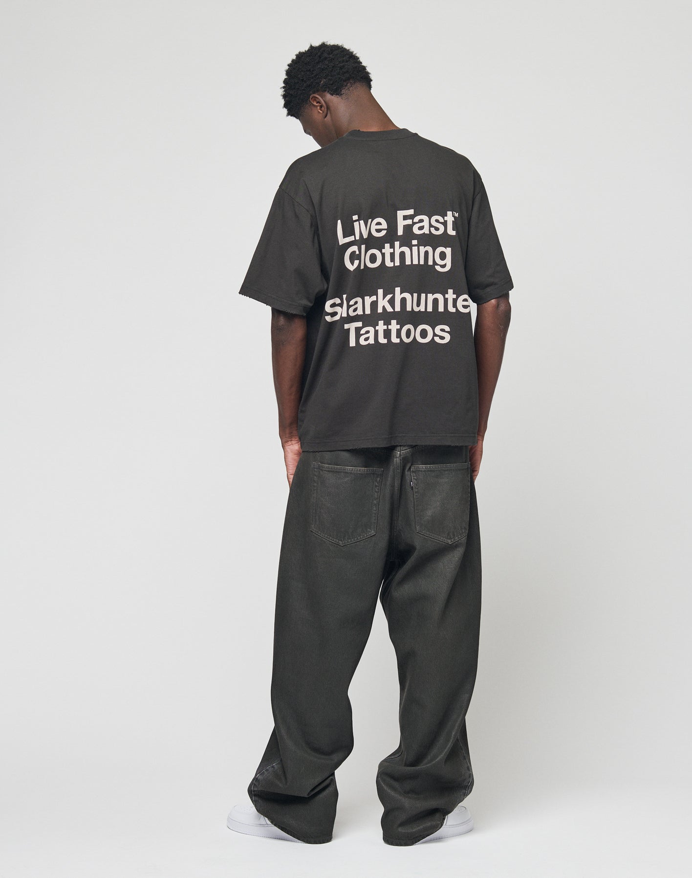 A person stands facing away, wearing an oversize black LFDY Sharkhunter Tee by LIVE FAST DIE YOUNG with white text and loose black jeans against a plain white background.