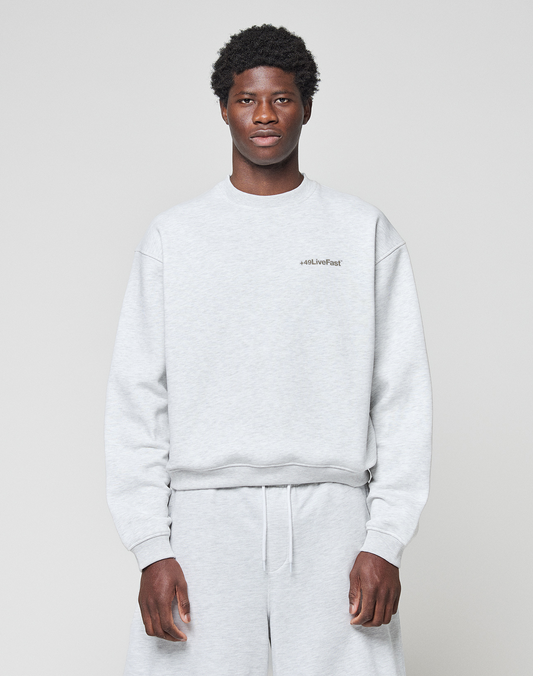 A young man stands against a plain light gray background wearing the LIVE FAST DIE YOUNG Basic +49 Chest Sweater and matching sweatpants, featuring a subtle chestprint near the left shoulder. He looks directly at the camera with a neutral expression. | model