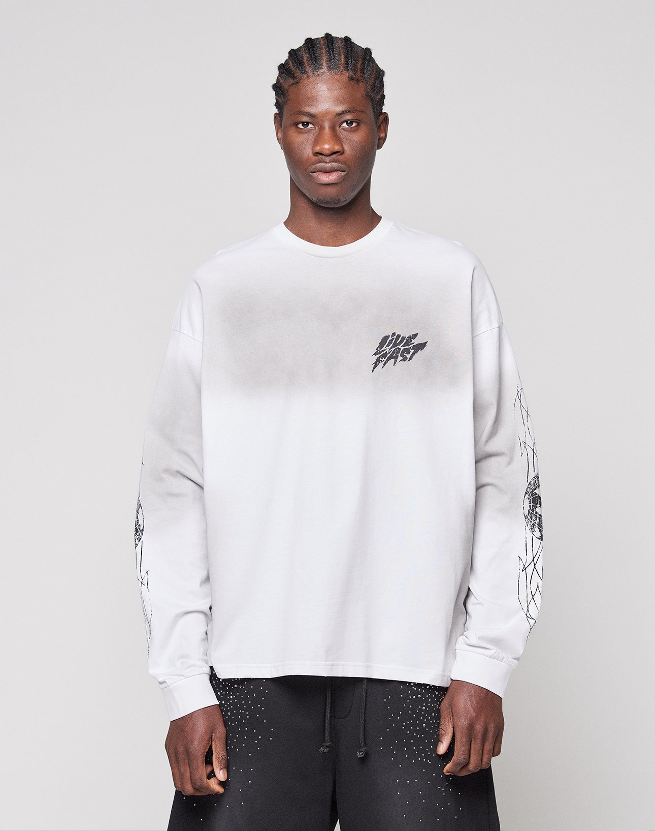 A man with cornrow braids wears the LIVE FAST DIE YOUNG LFDY Logo Longsleeve—an oversize, light gray 100% Baumwolle shirt with black graphics on chest and sleeves—paired with black speckled shorts, against a plain light background.