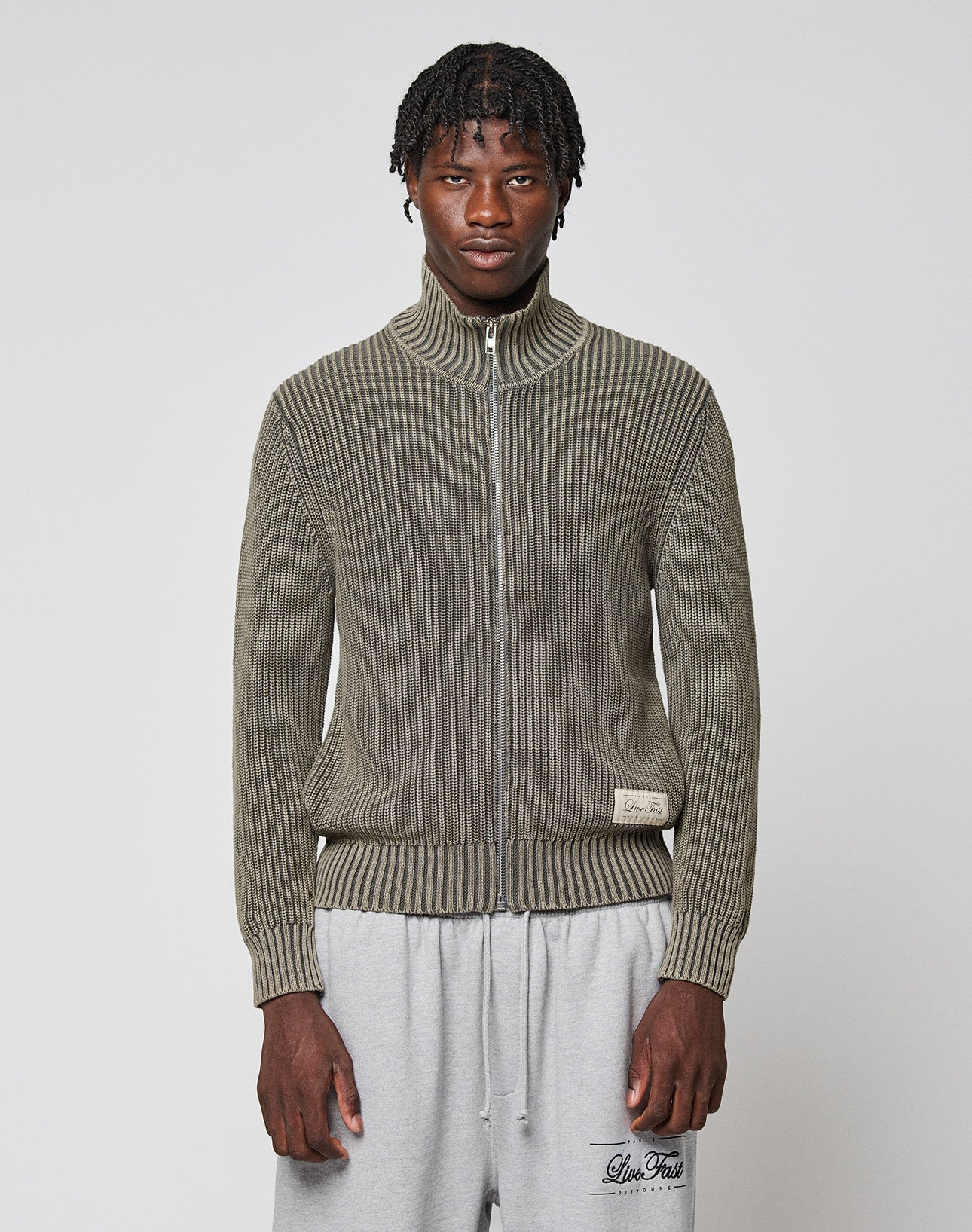 A man with short twisted hair wears the LFDY Knit Jacket from LIVE FAST DIE YOUNG in olive green and light gray sweatpants, standing against a plain white background. | model