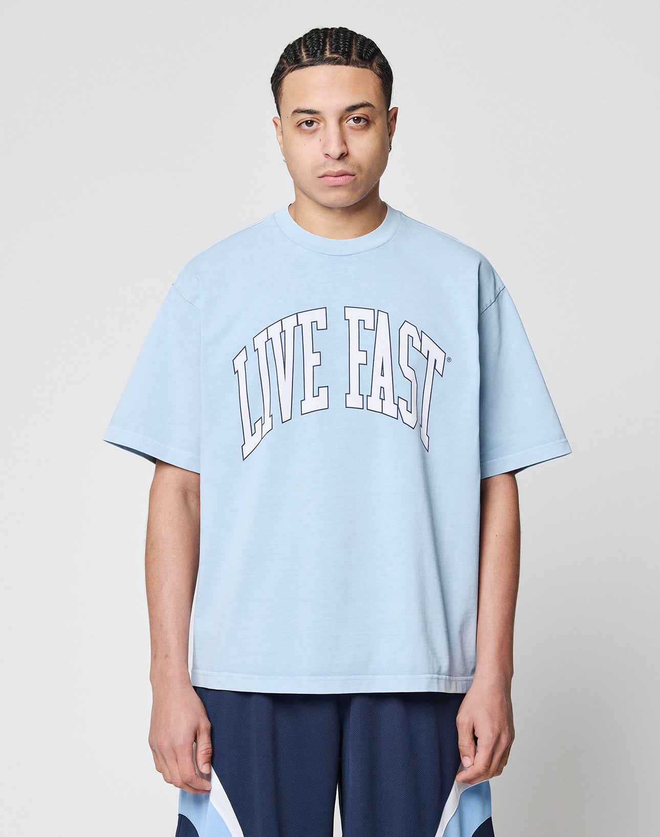 A person with braided hair wears the Live Fast College Tee by LIVE FAST DIE YOUNG—a light blue, 100% cotton oversized T-shirt with “LIVE FAST” in large white letters—standing against a plain background and looking at the camera.