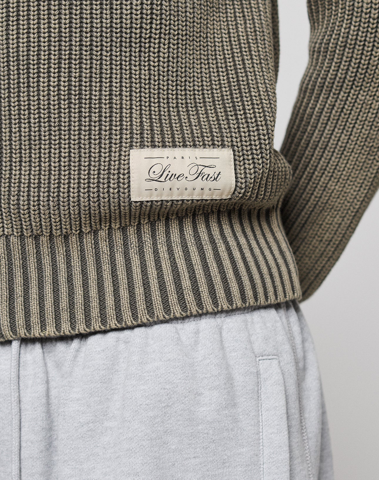 Close-up of the lower LFDY Knit Jacket by LIVE FAST DIE YOUNG in ribbed olive-green with a beige Live Fast patch, paired with light gray sweatpants. Only part of the jacket and upper pants are shown.