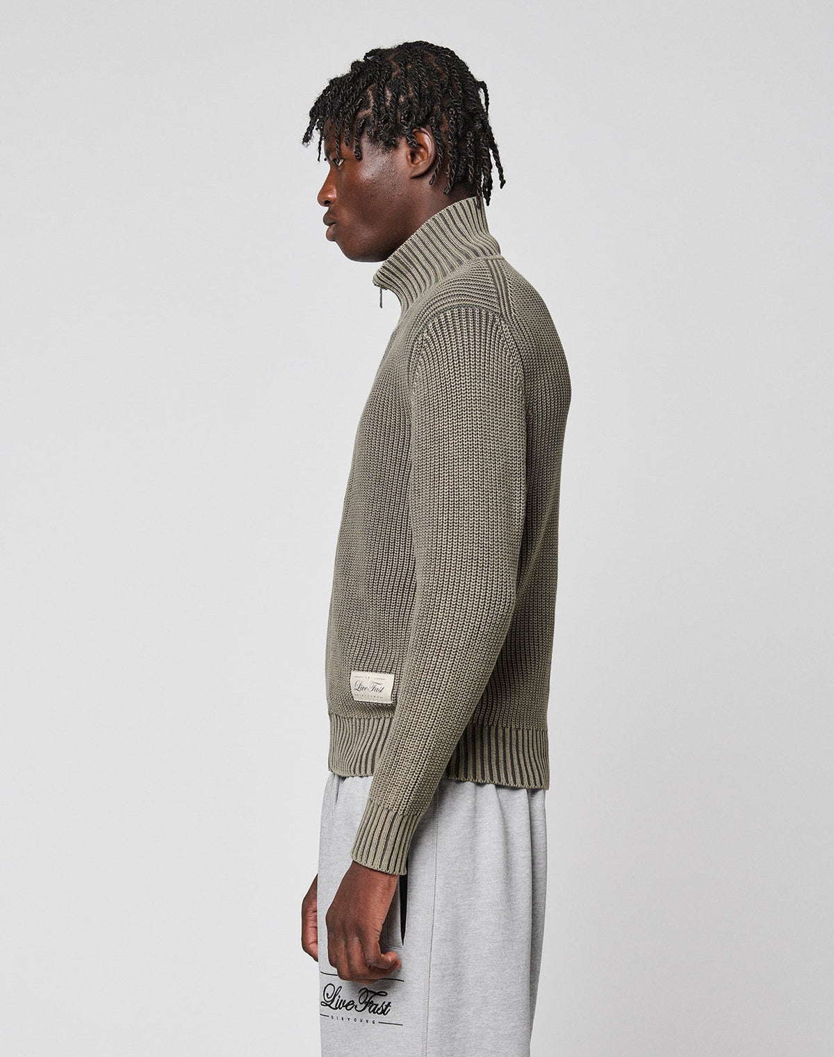 A person stands in profile wearing the LIVE FAST DIE YOUNG LFDY Knit Jacket with a stand-up collar and light gray sweatpants against a plain light gray background.