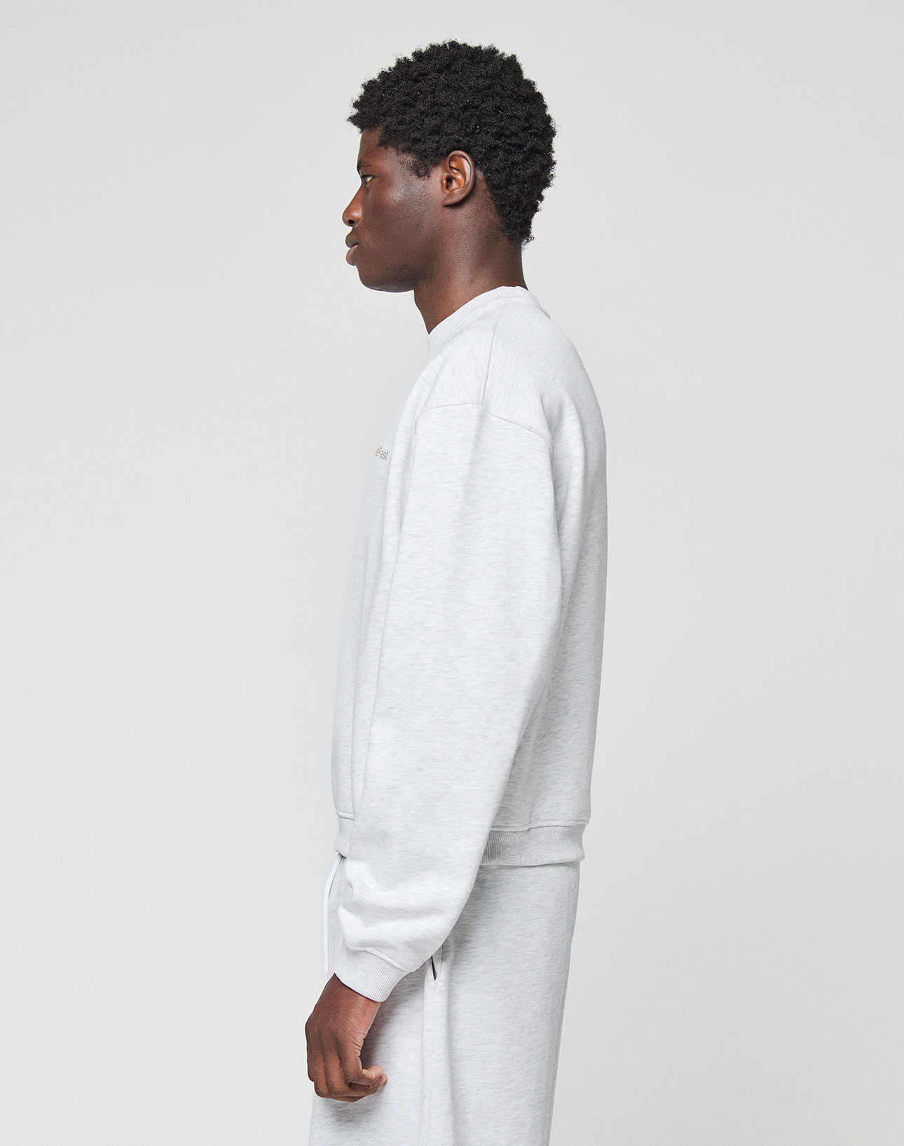 Someone stands in profile against a plain, light background wearing the LIVE FAST DIE YOUNG Basic +49 Chest Sweater and matching sweatpants in an oversize fit and light gray Baumwolle fabric.