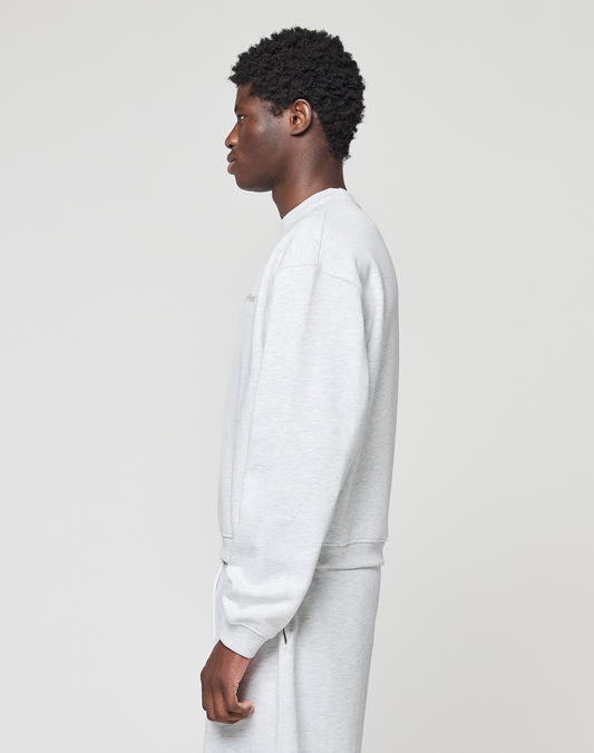 Someone stands in profile against a plain, light background wearing the LIVE FAST DIE YOUNG Basic +49 Chest Sweater and matching sweatpants in an oversize fit and light gray Baumwolle fabric.