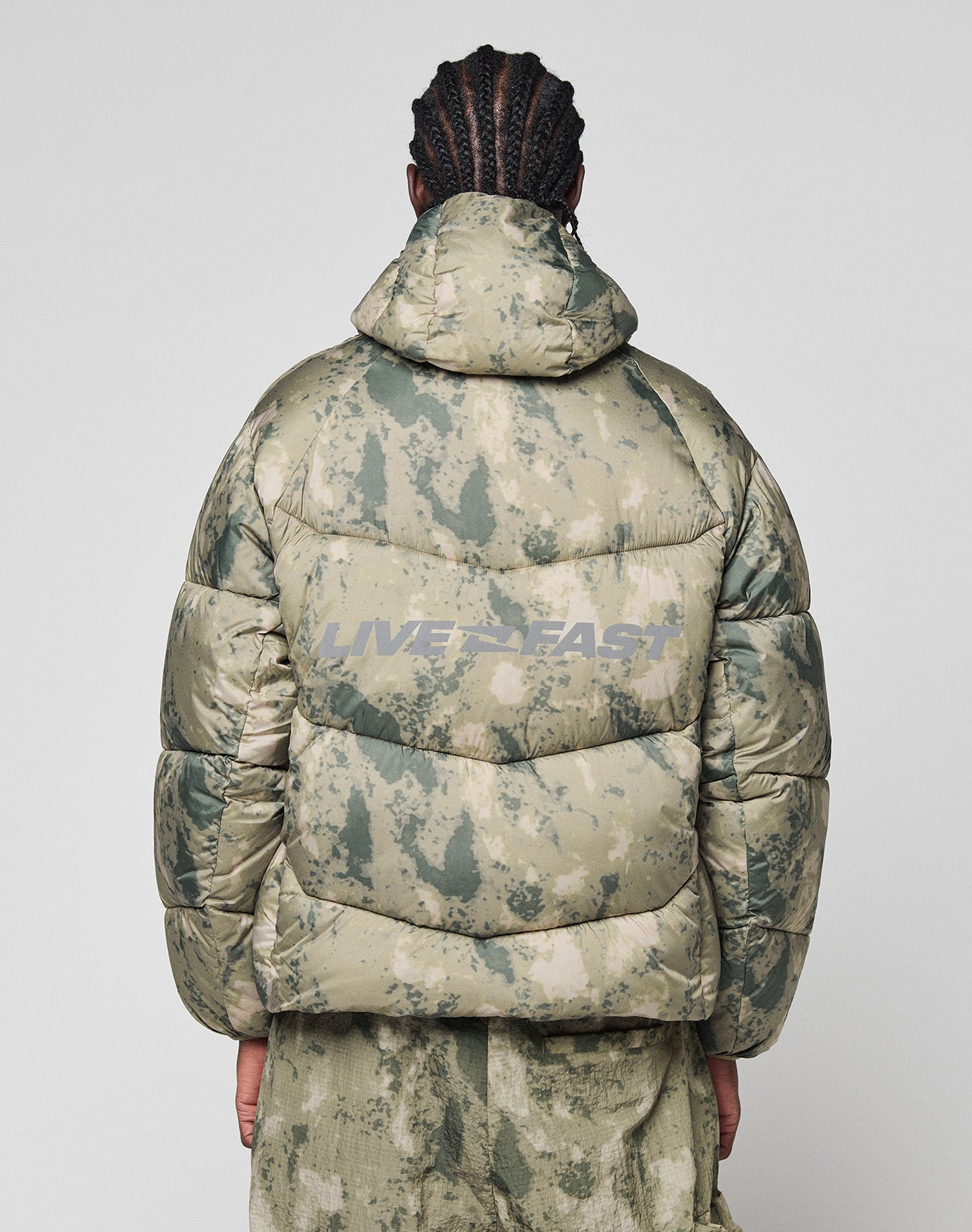 A person with braided hair, seen from behind, wears the LIVE FAST DIE YOUNG Live Fast Puffer Jacket—an oversized green camo jacket with hood and bold gray reflective "LIVE FAST" print on the back.