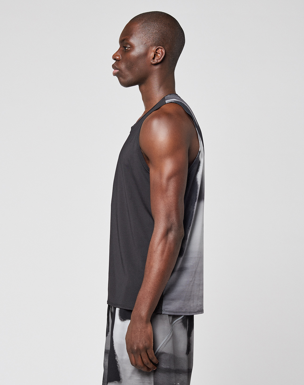 A man stands in profile against a plain background, wearing the LIVE FAST DIE YOUNG LFDY Performance Tank with a black and gray tie-dye design, reflective logo print, matching lightweight shorts, and Regular Fit for a sporty look.
