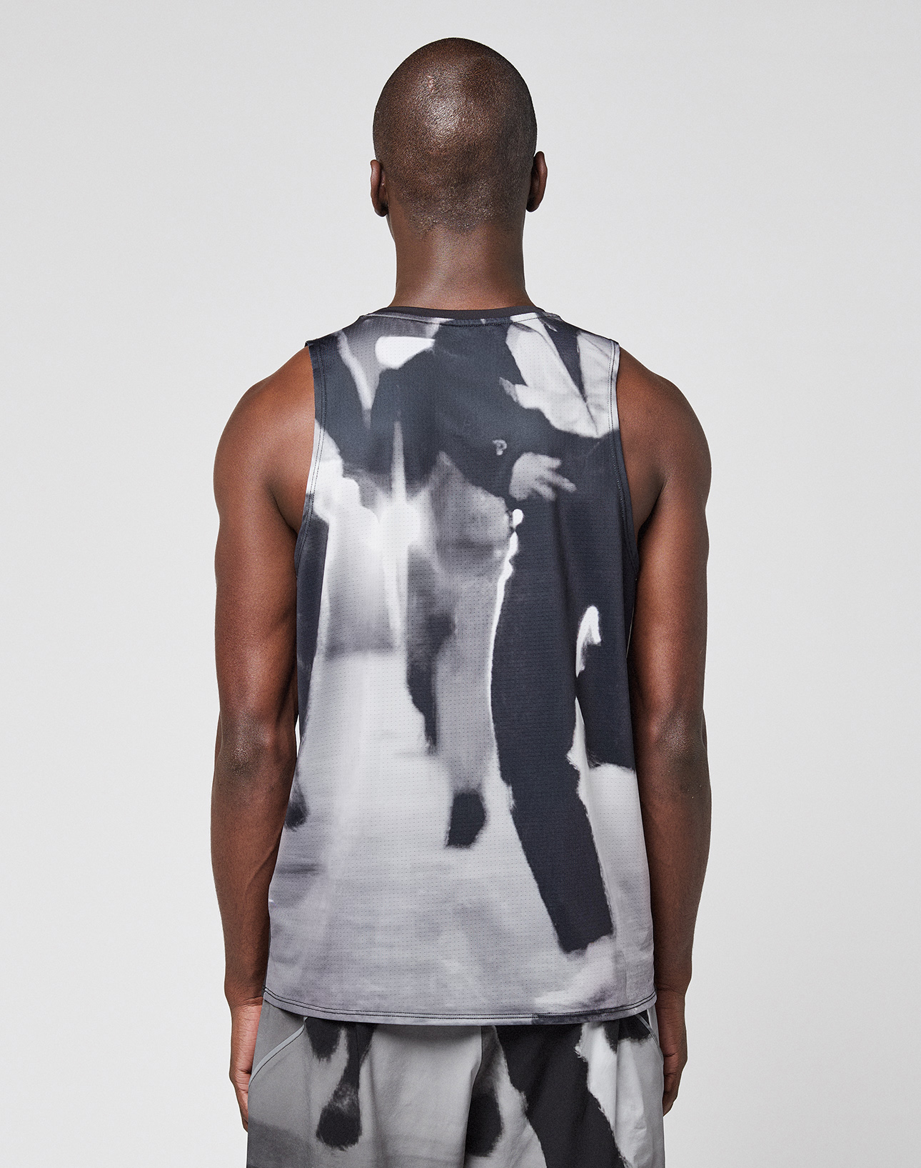 Seen from behind, a person with a shaved head wears the LFDY Performance Tank by LIVE FAST DIE YOUNG, made from leichtes Obermaterial with a black-and-white blurred print of people in suits. The Regular Fit stands out against a plain light gray background. | model