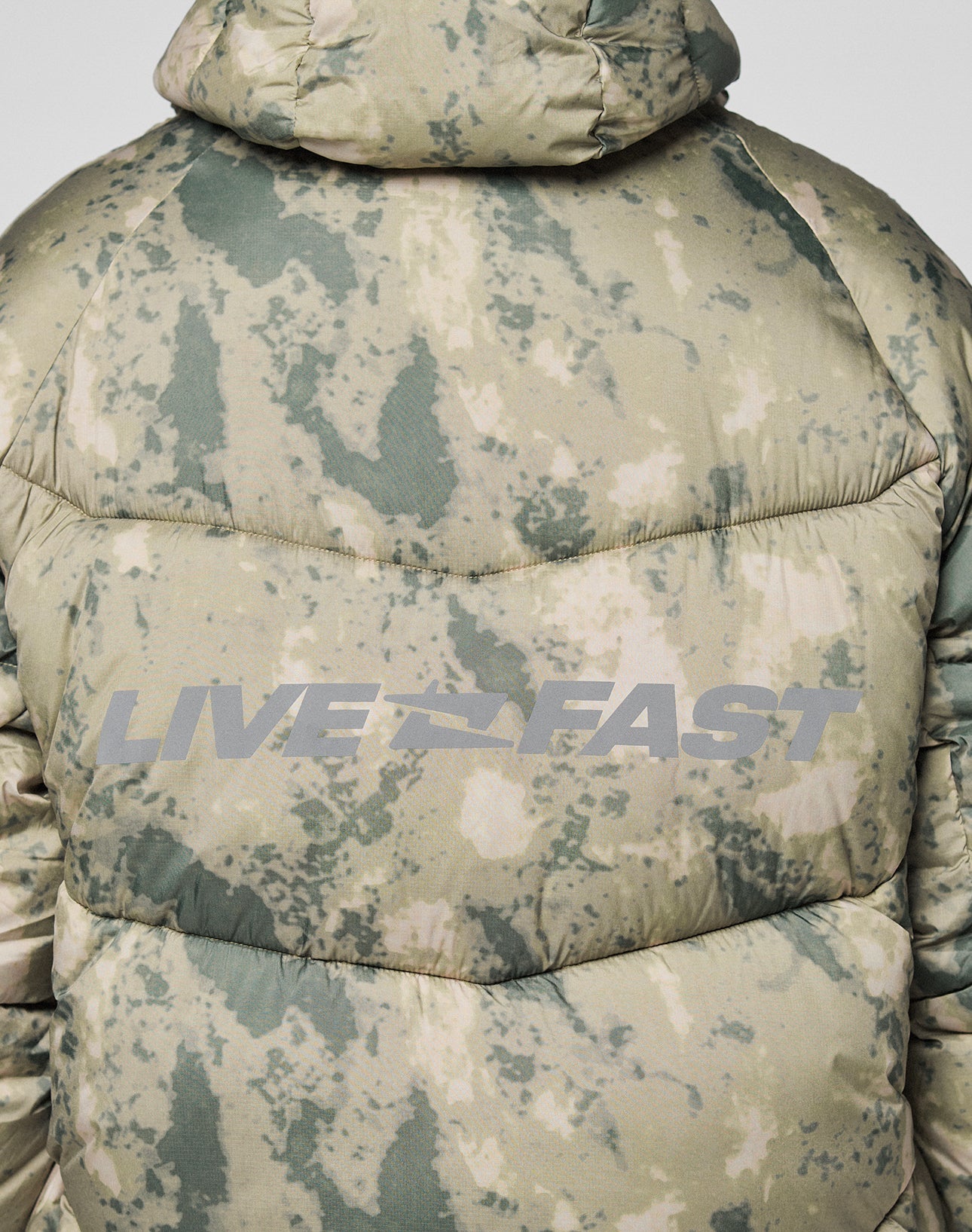 A close-up of the back of the green and beige Live Fast Puffer Jacket by LIVE FAST DIE YOUNG, featuring an oversized fit and large reflective "LIVE FAST" lettering across the back.