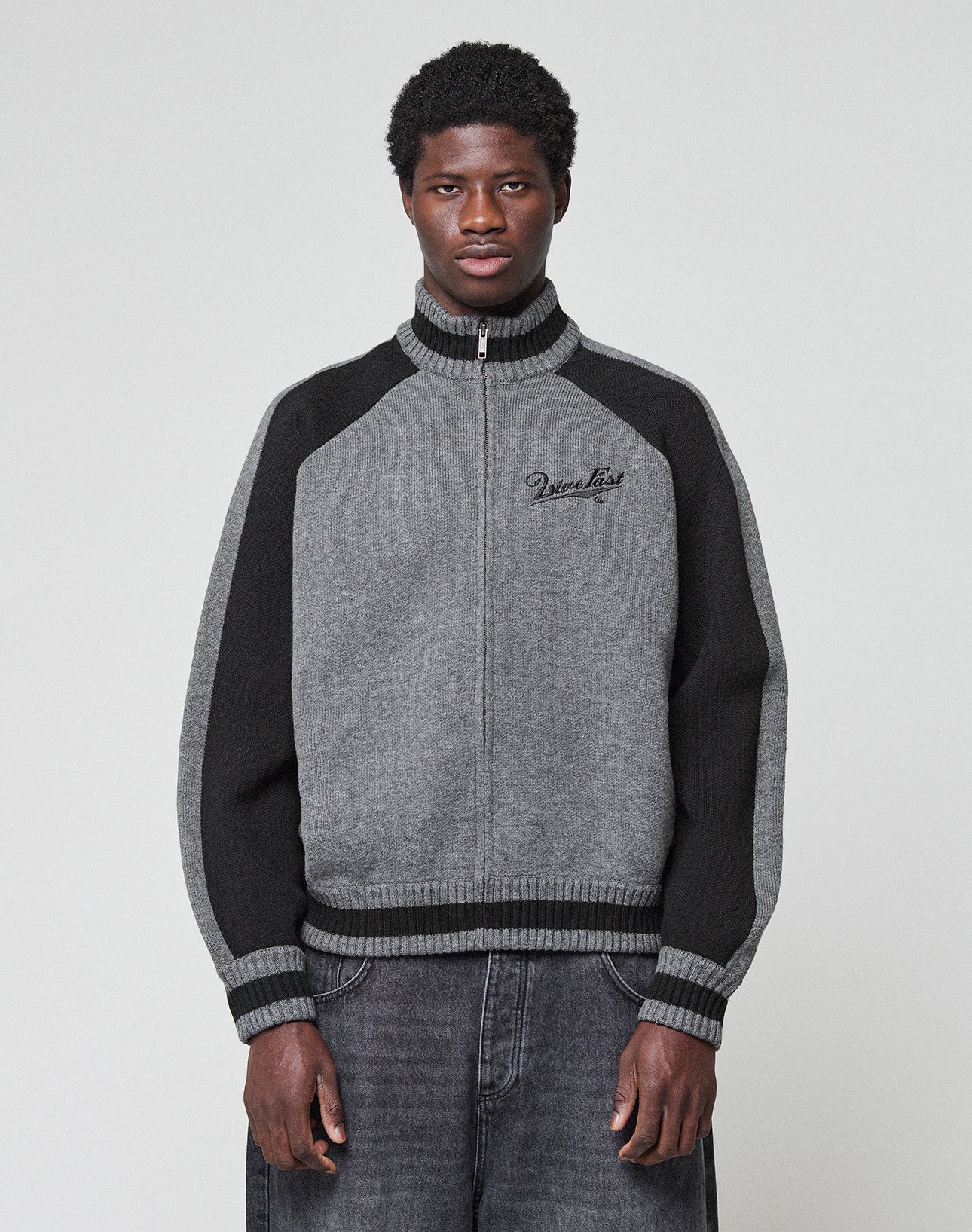 The LIVE FAST DIE YOUNG Boxy Knit Zip Jacket features gray and black intarsia-knit, raglan sleeves, ribbed collar, cuffs, and hem, black side panels, two front pockets, and embroidered cursive text on the left chest.