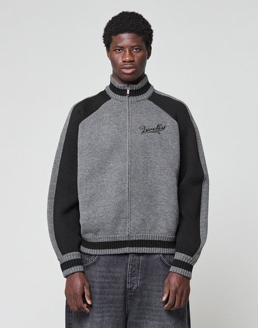 A man with short curly hair faces forward in a LIVE FAST DIE YOUNG Boxy Knit Zip Jacket, oversize fit, grey and black with raglan sleeves and a Vinculer logo on the chest, paired with loose grey jeans against a plain light background.
