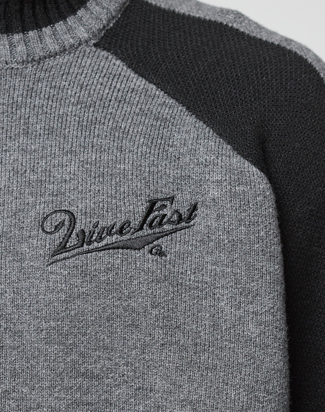 A man with short curly hair faces forward in a LIVE FAST DIE YOUNG Boxy Knit Zip Jacket, oversize fit, grey and black with raglan sleeves and a Vinculer logo on the chest, paired with loose grey jeans against a plain light background.