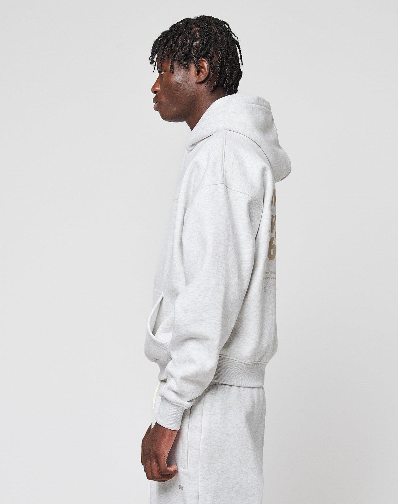 A person with short dreadlocks stands in profile against a plain light background, wearing the LIVE FAST DIE YOUNG Basic 360 Hooded sweatshirt and matching sweatpants in light gray Oversize Fit.