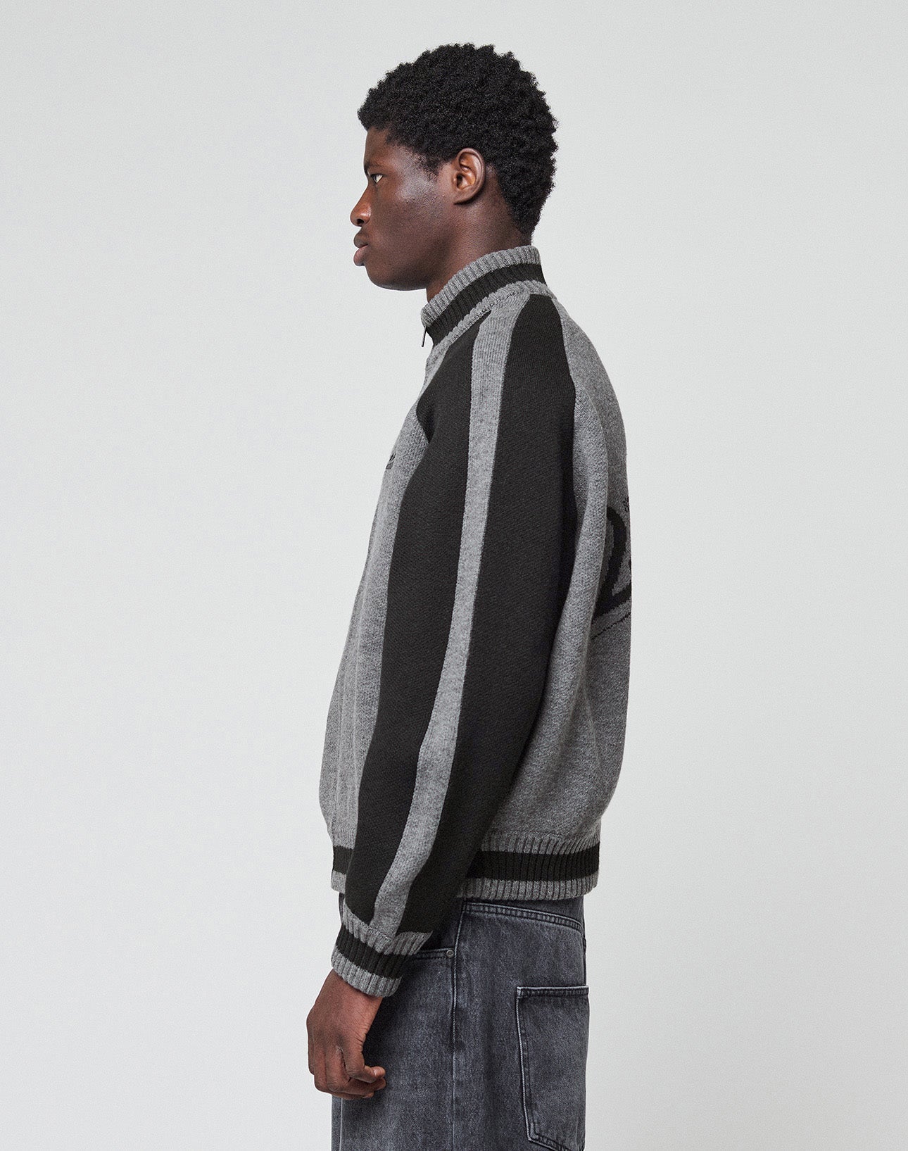 A person with short curly hair wears the LIVE FAST DIE YOUNG Boxy Knit Zip Jacket in gray and black with a galloping horse intarsia and “Live Fast Co.” on the back, paired with faded gray jeans, standing facing away from the camera.| model