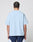 A person with neatly braided hair is shown from the back, wearing the LIVE FAST DIE YOUNG Live Fast College Tee in oversize light blue 100% Baumwolle and navy shorts with white accents, standing against a plain light gray background.