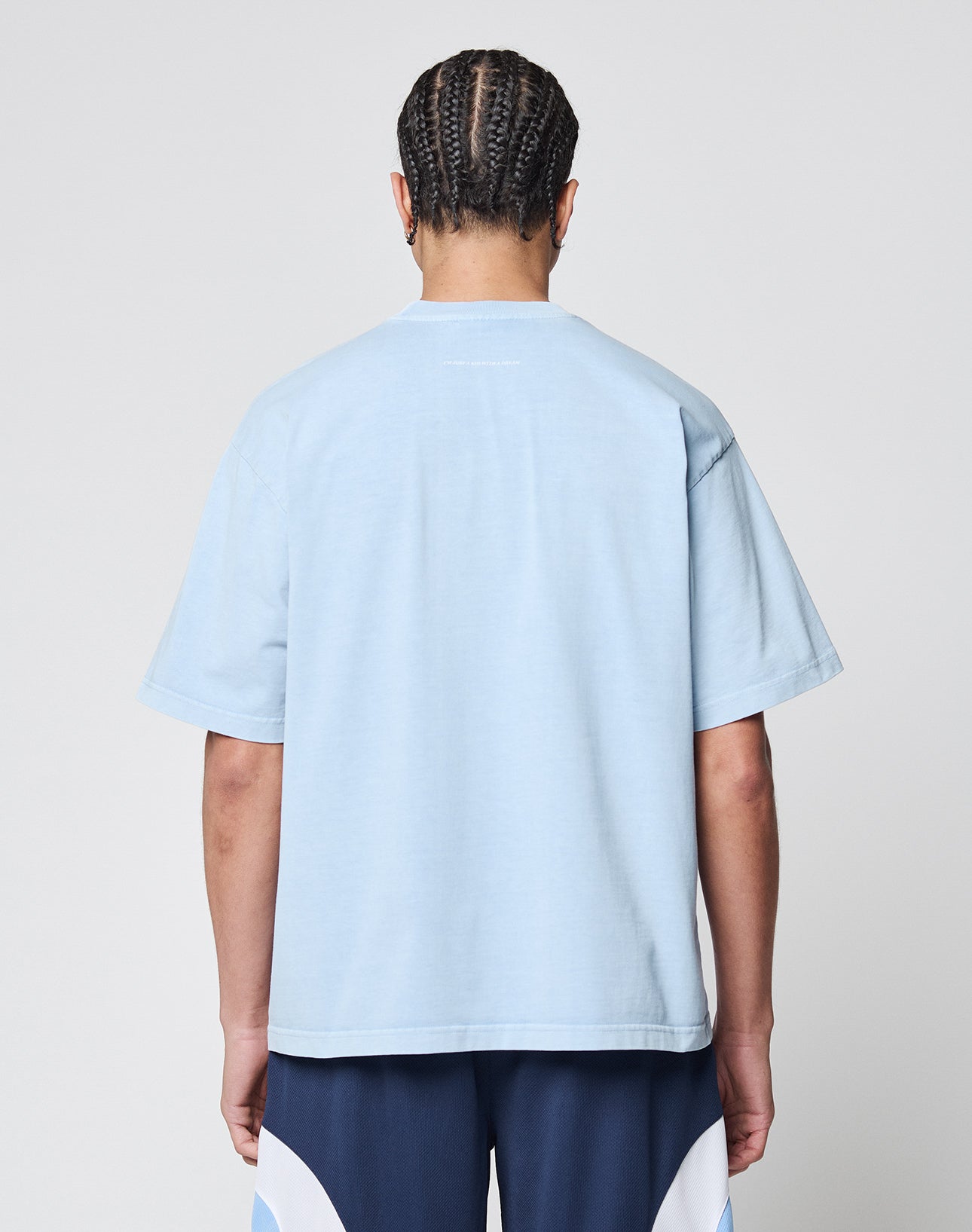 A person with neatly braided hair is shown from the back, wearing the LIVE FAST DIE YOUNG Live Fast College Tee in oversize light blue 100% Baumwolle and navy shorts with white accents, standing against a plain light gray background.