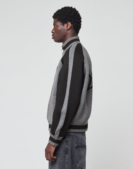 A person stands in profile against a plain background, wearing the LIVE FAST DIE YOUNG Boxy Knit Zip Jacket with gray and black vertical-striped sleeves, paired with dark gray jeans.