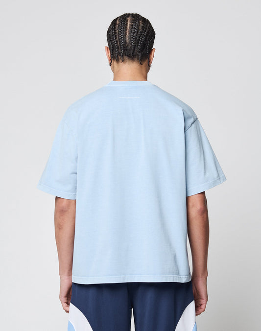 A person with neatly braided hair is shown from the back, wearing the LIVE FAST DIE YOUNG Live Fast College Tee in oversize light blue 100% Baumwolle and navy shorts with white accents, standing against a plain light gray background.