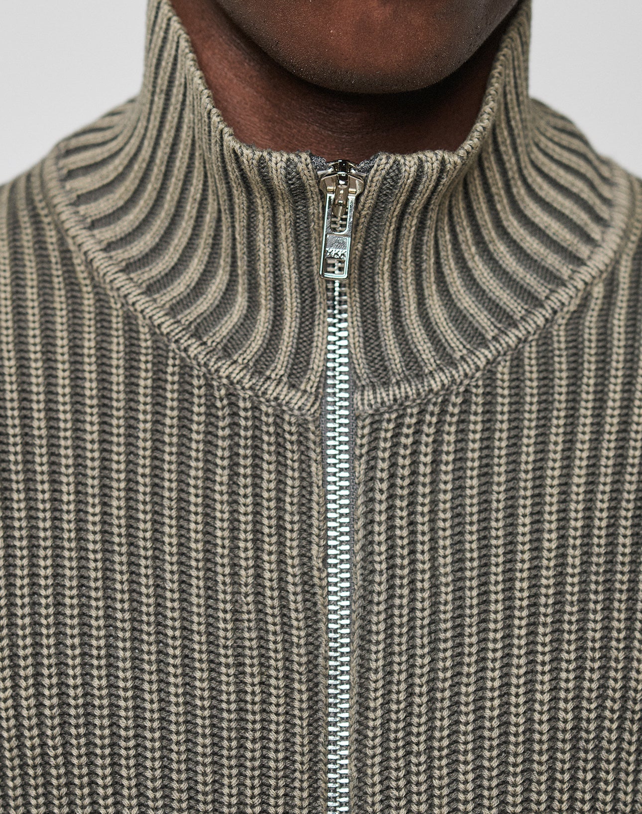 Close-up of a person wearing the LFDY Knit Jacket by LIVE FAST DIE YOUNG, featuring ribbed texture, high collar, and zip-up design in gray and beige shades. Zipper is partially closed; only the lower half of the face is visible.