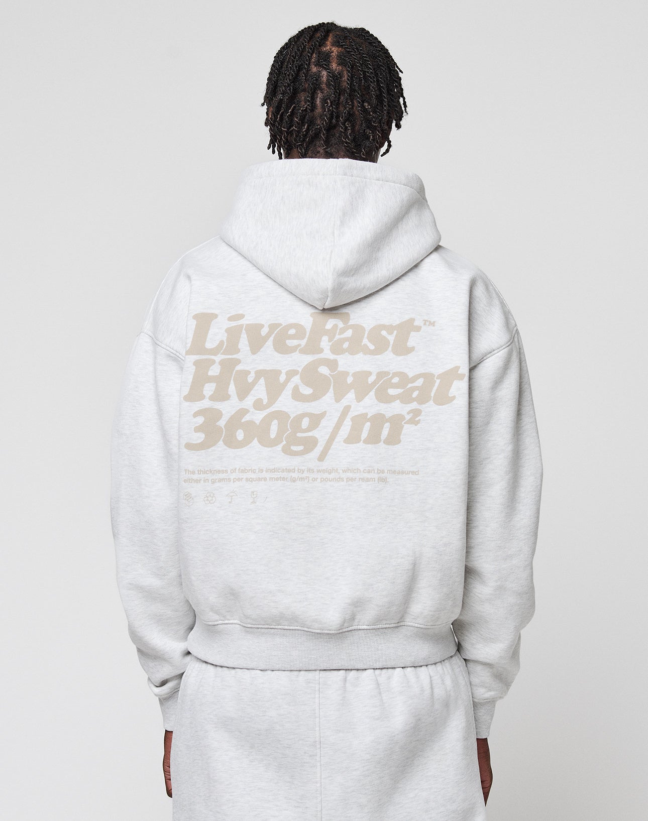 A person with braided hair is seen from the back wearing an oversize fit light gray Basic 360 Hooded and sweatpants by LIVE FAST DIE YOUNG, featuring a bold beige “Live Fast Hyv Sweat 360g/m²” backprint.