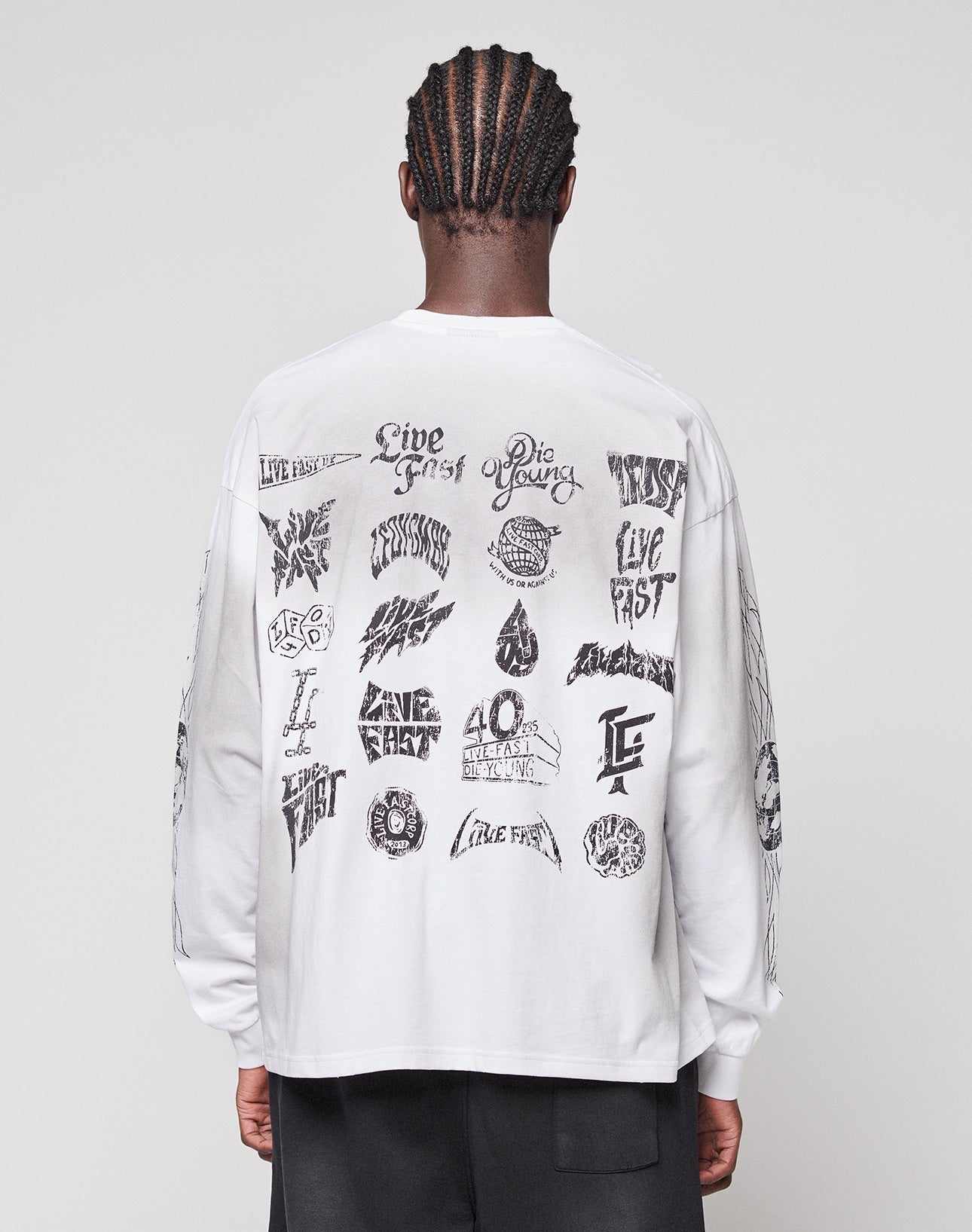 A person with braided hair faces away, wearing the LIVE FAST DIE YOUNG LFDY Logo Longsleeve in oversize fit, featuring black graphic designs and logos across the back and sleeves.