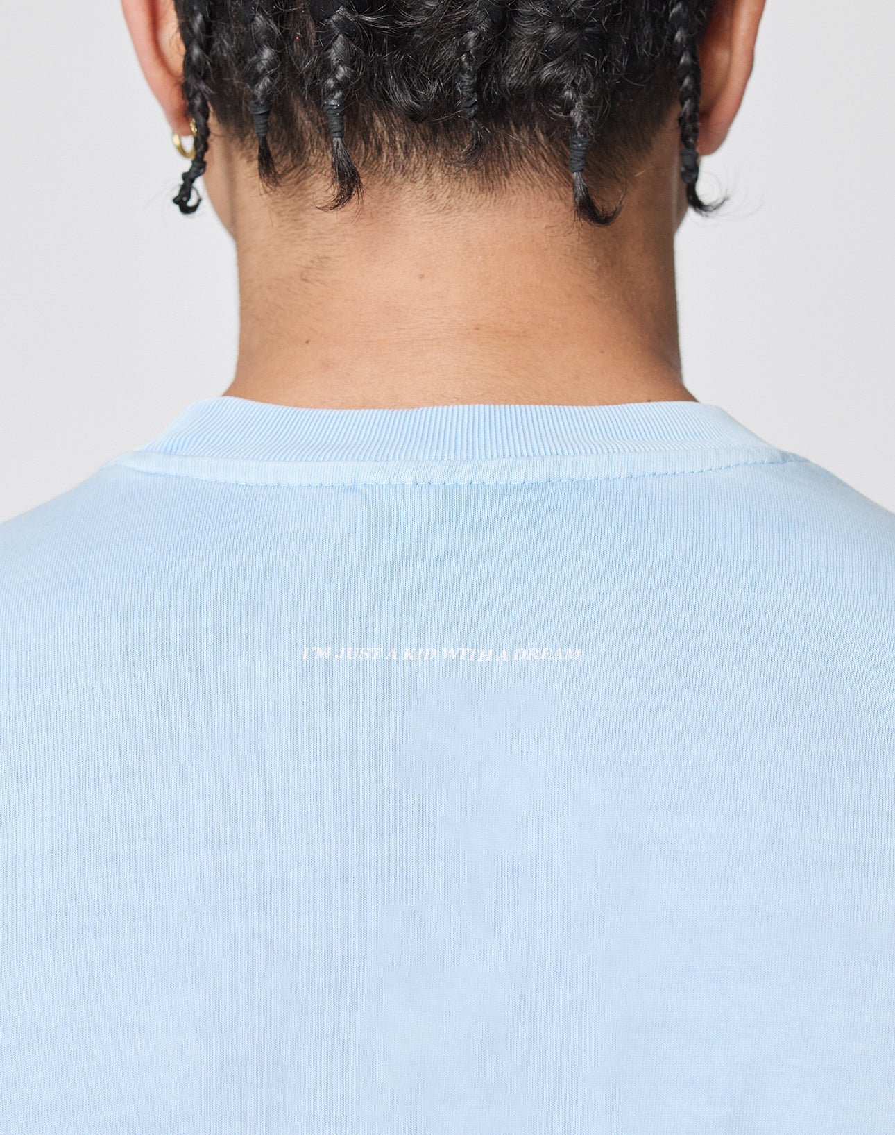 The back of a person’s head with braided hair, wearing the Live Fast College Tee by LIVE FAST DIE YOUNG in Oversize Fit. Small white text near the collar reads, “I’M JUST A KID WITH A DREAM.”.