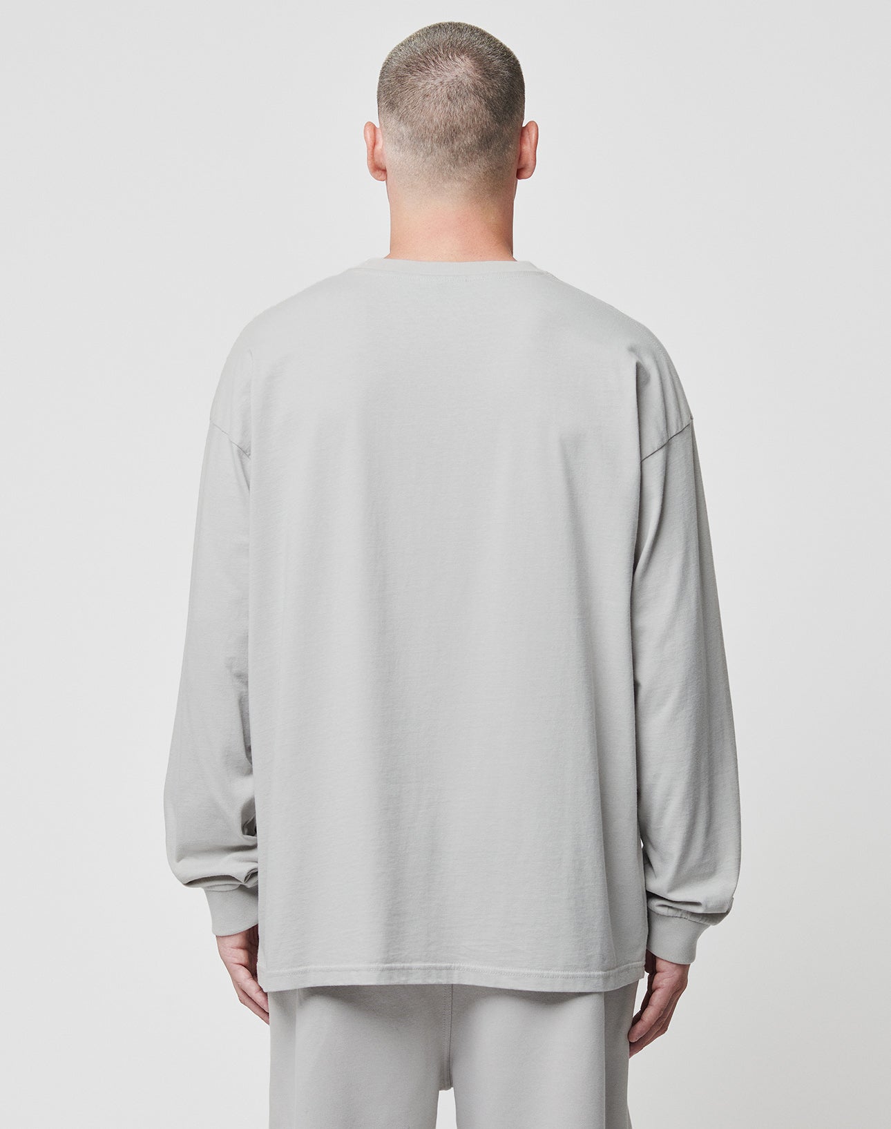 A person with short hair, seen from behind, wears the LIVE FAST DIE YOUNG Basic Longsleeve in an oversize fit and light gray 100% Baumwolle fabric with matching pants against a plain white background.