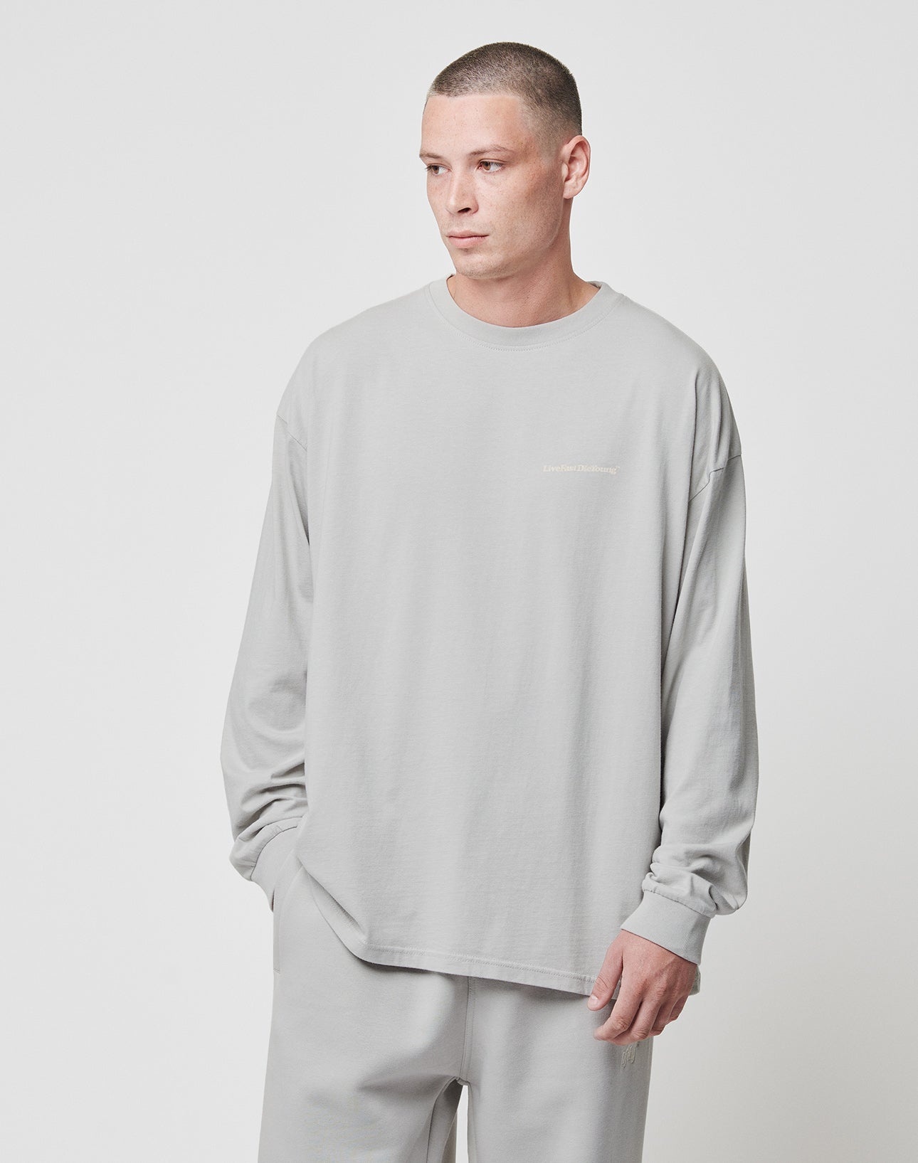 A person with short hair wears the LIVE FAST DIE YOUNG Basic Longsleeve and matching pants in light gray 100% Baumwolle, standing against a plain white background and looking slightly to the side.