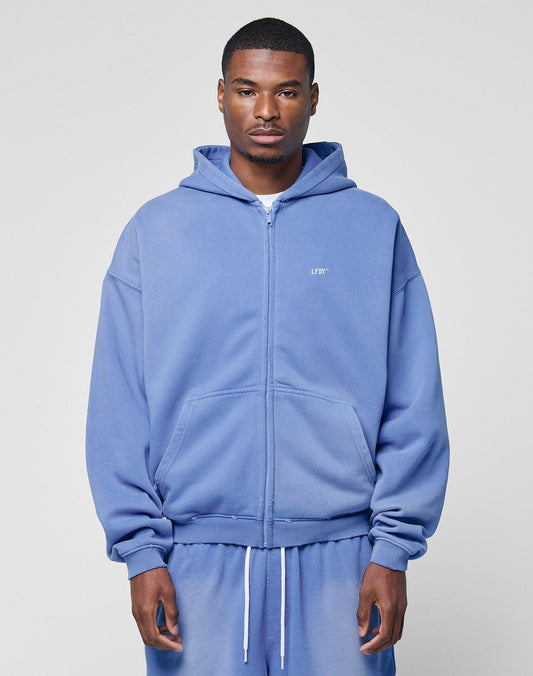 A man wears the LIVE FAST DIE YOUNG Basic Hooded Zip in light blue with Kapuze, front pockets, and handgefertigtem Fade-Effekt, standing before a plain light gray background and looking neutrally at the camera.