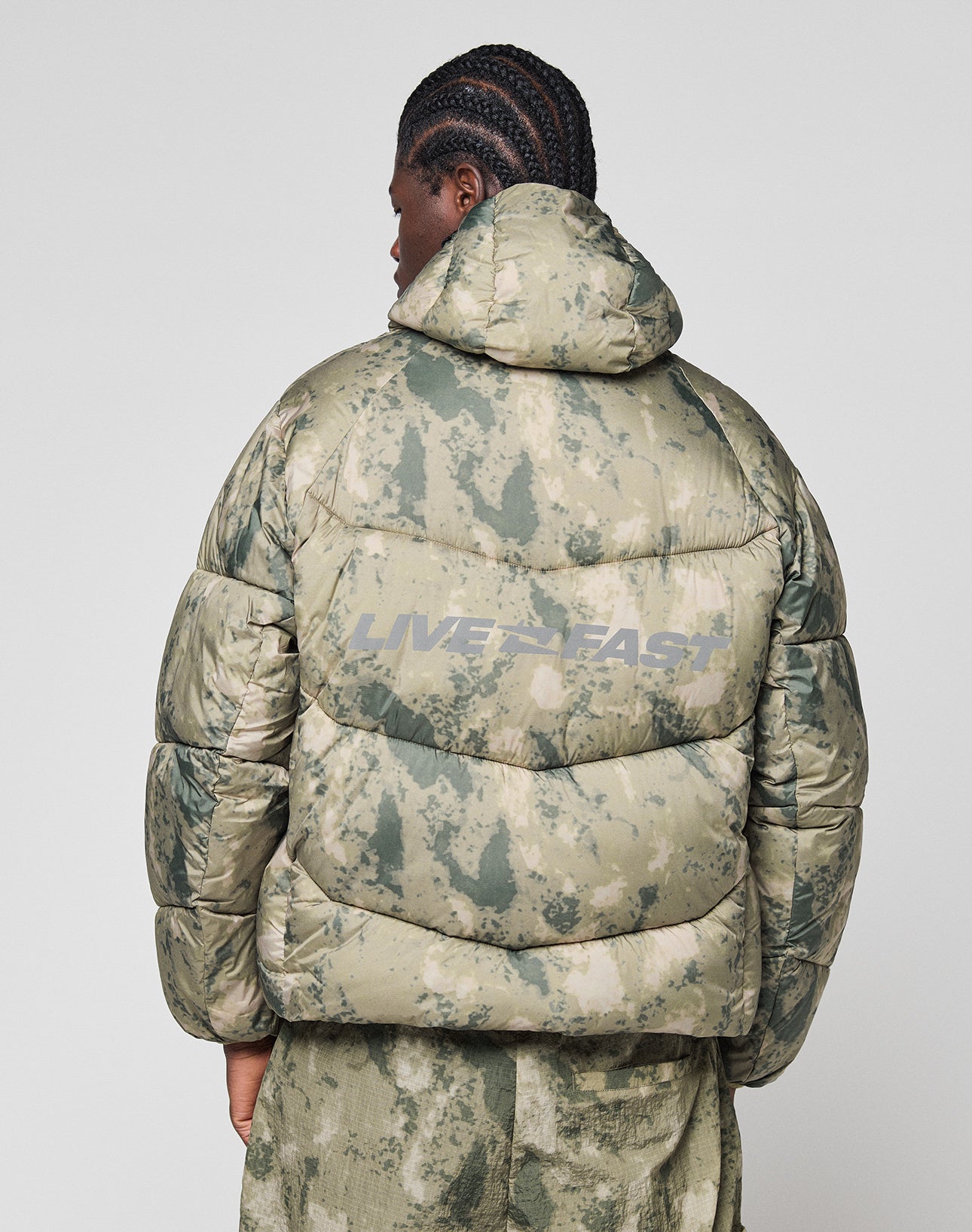 A person with braided hair is seen from behind wearing the LIVE FAST DIE YOUNG Live Fast Puffer Jacket, an oversized green and beige camo style featuring “LIVE FAST” in large reflective print on the back.