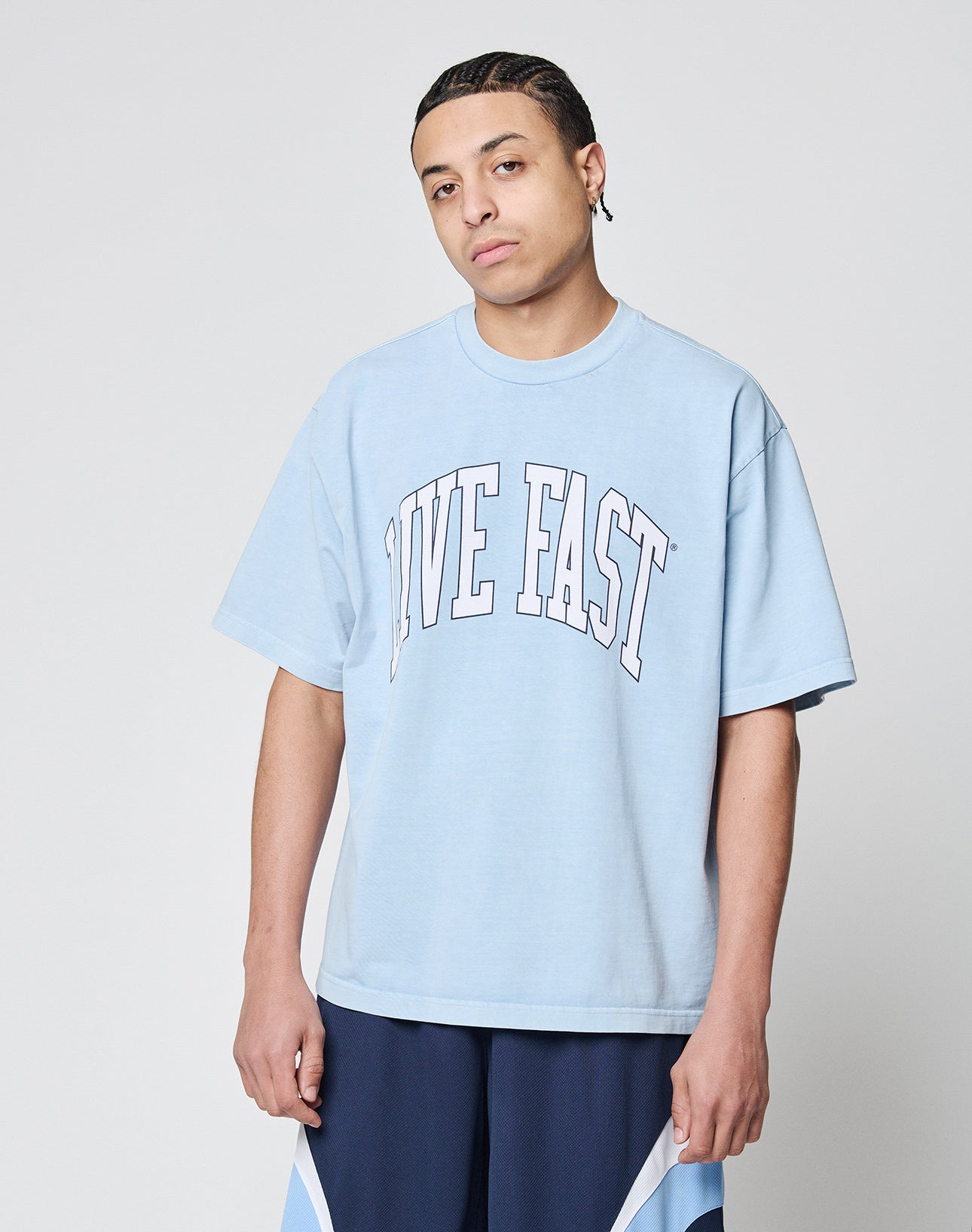 A person with braided hair wears the LIVE FAST DIE YOUNG Live Fast College Tee in light blue Oversize Fit, featuring LIVE FAST in bold white letters, paired with blue and white shorts, standing against a plain light gray background.