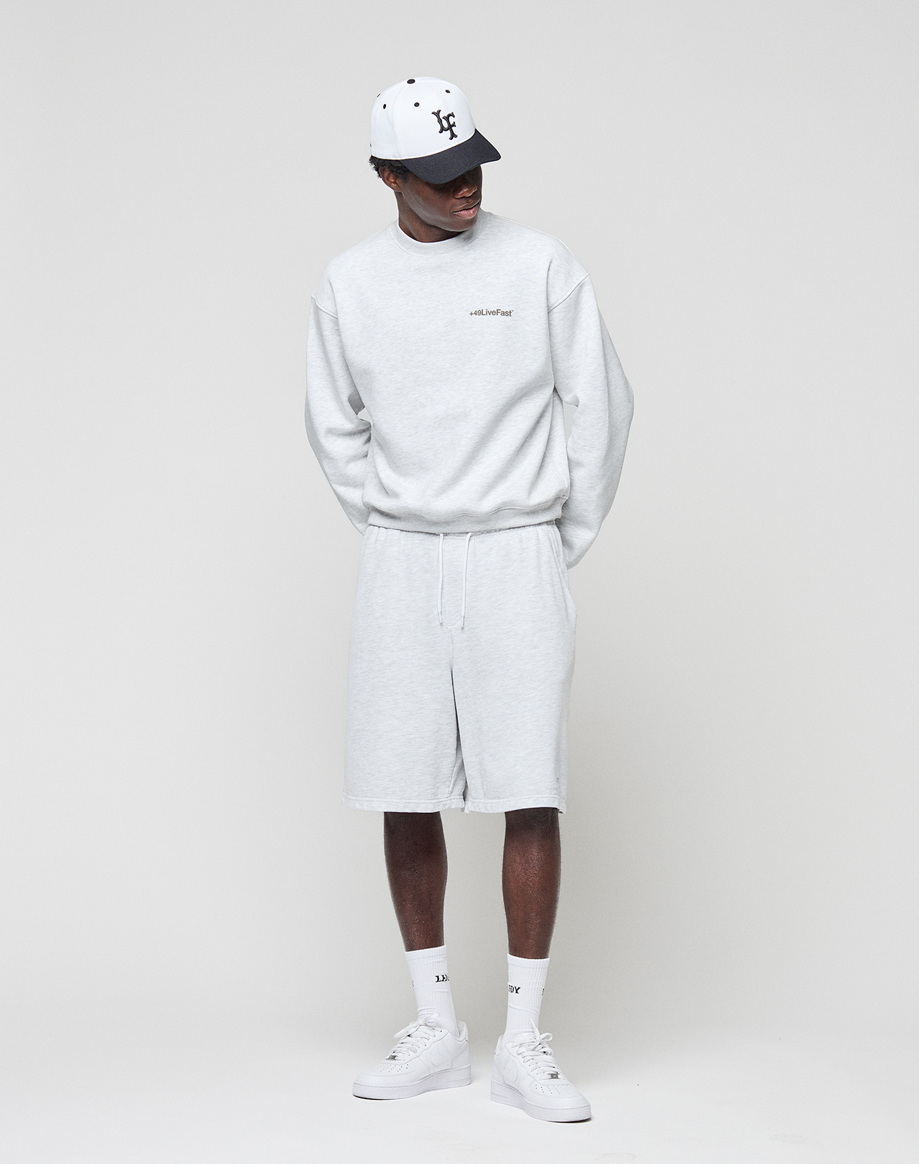 Wearing the LIVE FAST DIE YOUNG Basic +49 Chest Sweater in light grey and matching shorts, white socks, white sneakers, and a white baseball cap, a person stands against a plain background looking to the side with hands behind their back.