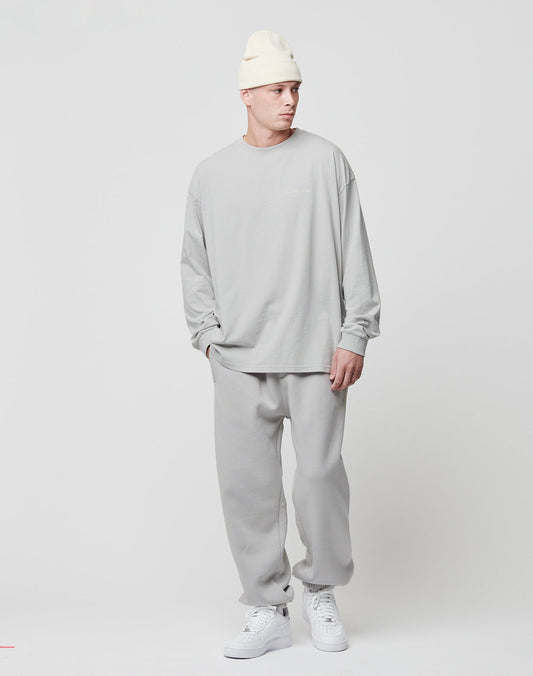 A person stands against a plain white background wearing the LIVE FAST DIE YOUNG Basic Longsleeve in light gray with matching sweatpants, white sneakers, and a cream beanie. One hand is in their pocket as they look to the side.