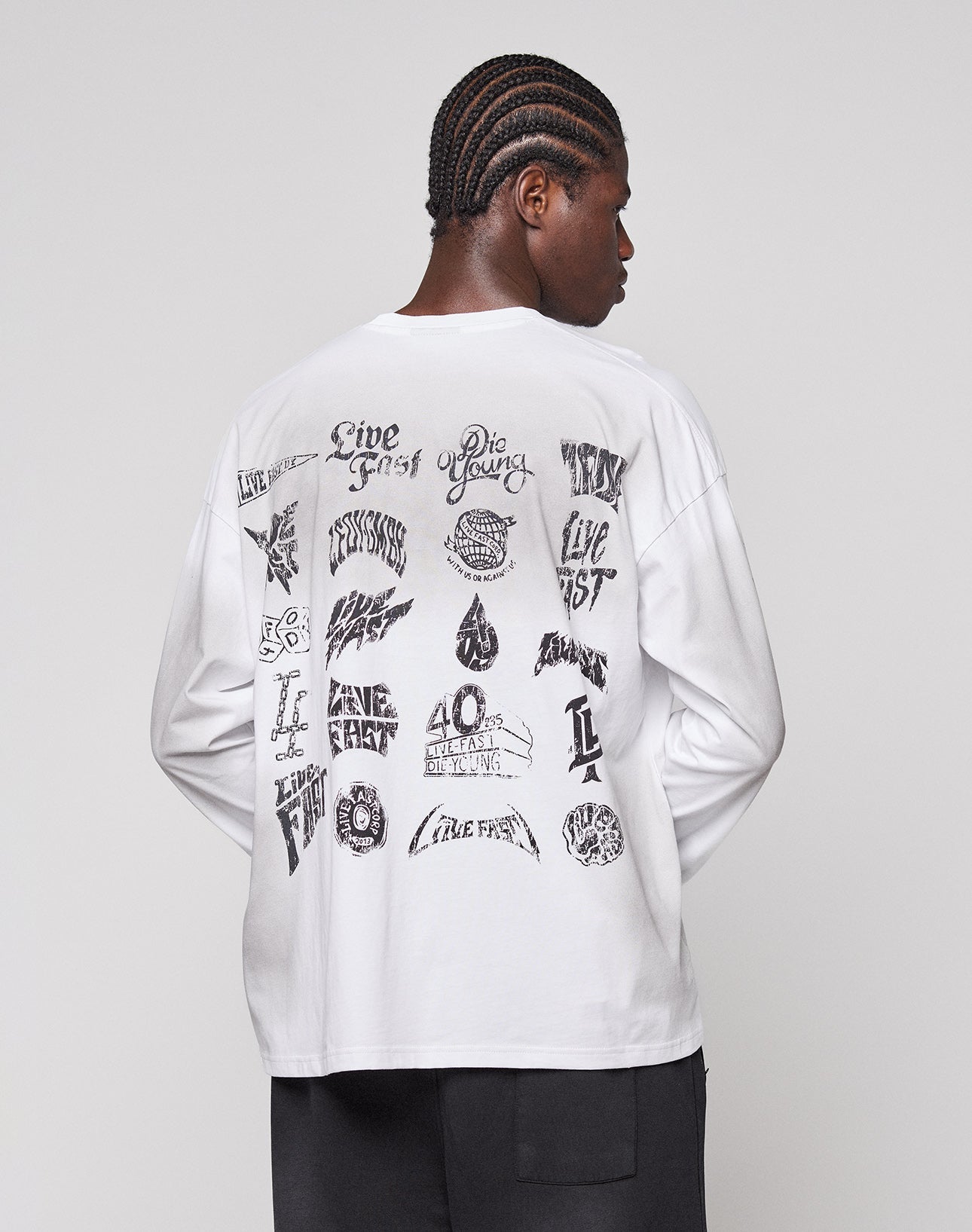 A person with braided hair wears the LFDY Logo Longsleeve by LIVE FAST DIE YOUNG, a white oversized 100% Baumwolle shirt with black graphic designs and text on the back, standing against a plain light gray background.