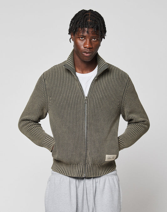 A person with short, braided hair wears an olive-green LFDY Knit Jacket by LIVE FAST DIE YOUNG over a white t-shirt and light gray sweatpants, standing against a plain light gray background.