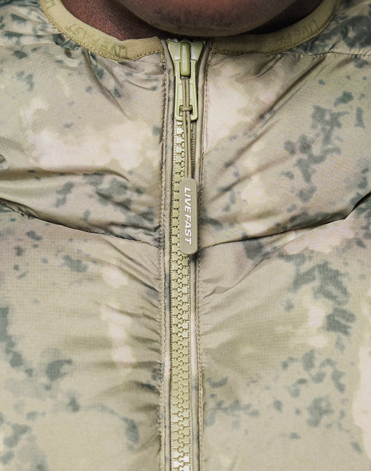 Close-up of the Live Fast Puffer Jacket by LIVE FAST DIE YOUNG, featuring a light camo pattern in green and beige, a zipper pull printed with LIVE FAST, and wasserabweisendes material for extra protection.