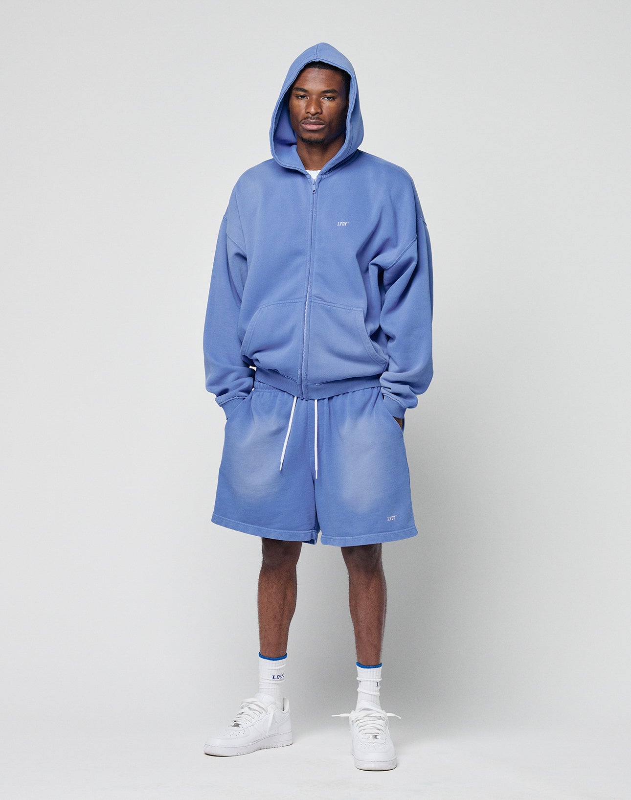 A man faces forward in a plain light gray setting, wearing LIVE FAST DIE YOUNG Basic Sweat Shorts and a matching blue hoodie with the hood up and zipped, paired with white socks and sneakers. The shorts have an elastic waistband.