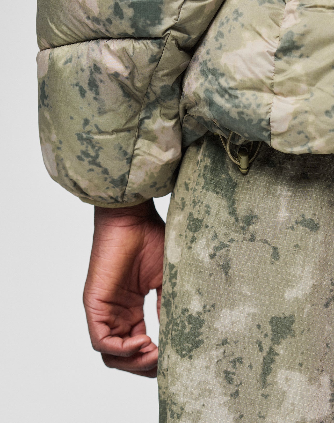 A close-up of a hand and arm wearing the LIVE FAST DIE YOUNG Live Fast Puffer Jacket in an oversize green camo print with matching pants and visible drawstring, set against a plain light background.