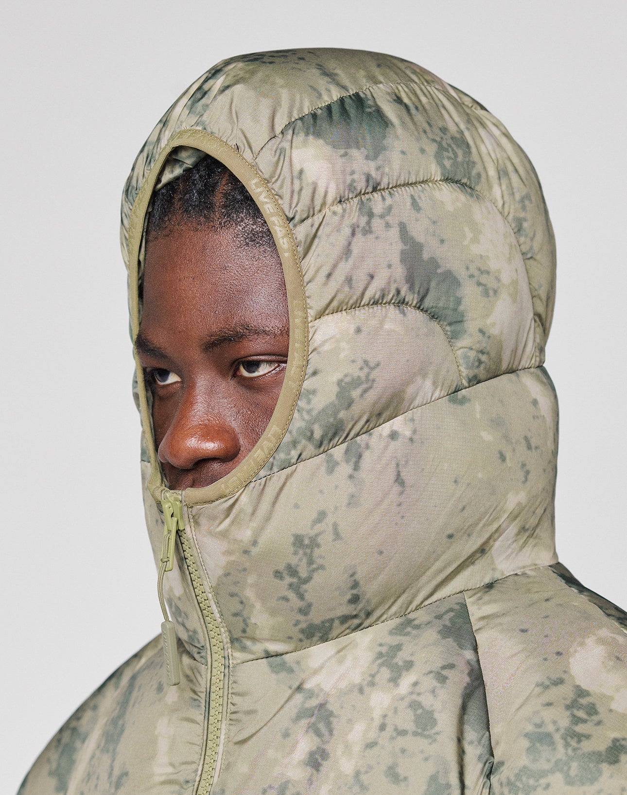 Someone wearing the LIVE FAST DIE YOUNG Live Fast Puffer Jacket in green and beige camo with an Oversize Fit and hood fully zipped, only their eyes visible, stands against a plain light background.