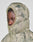 Someone wearing the LIVE FAST DIE YOUNG Live Fast Puffer Jacket in green and beige camo with an Oversize Fit and hood fully zipped, only their eyes visible, stands against a plain light background.