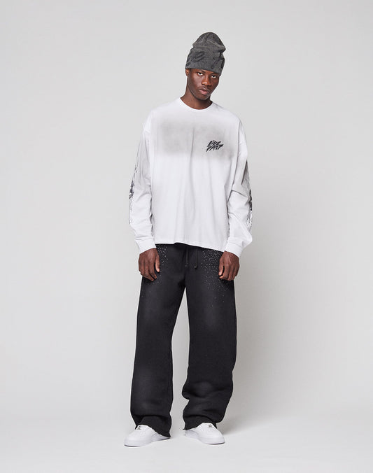 A man in an oversize gray beanie, loose black pants, white sneakers, and a LIVE FAST DIE YOUNG LFDY Logo Longsleeve with black graphics stands against a plain light background, looking at the camera with a neutral expression.