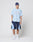 A person stands against a plain background wearing a blue headwrap, light blue LIVE FAST DIE YOUNG Oversize Fit T-shirt, navy and light blue Basic Trackshorts with elastischer Bund, white socks, and white sneakers. Hands in pockets.