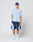 A person stands against a plain background wearing a blue headwrap, LIVE FAST DIE YOUNG’s Live Fast College Tee in light blue with LIVE FAST printed on it, dark blue athletic shorts with light blue accents, white socks, and white sneakers.