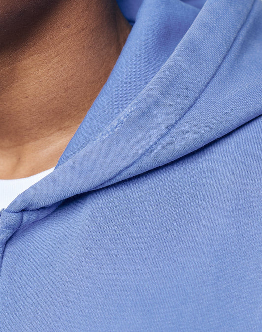 Close-up of a person wearing the LIVE FAST DIE YOUNG Basic Hooded Zip in light blue over a white shirt, showing the textured fabric, hood seam, and subtle distressing for a faded look. The edge of their face and neck is partially visible.