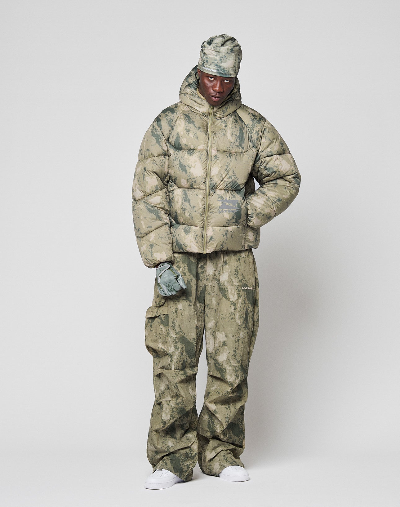 Wearing the LIVE FAST DIE YOUNG Live Fast Puffer Jacket in an oversized camo fit, cargo pants, gloves, headwrap, and white sneakers, a person stands against a white background. A reflektierender Print adds standout detail as they pose with one hand in a pocket.